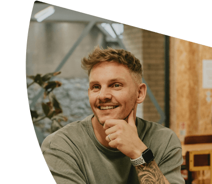 Smiling man with blonde hair and tattoos, thoughtfully looking at the viewer.