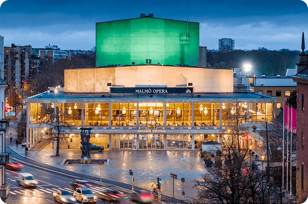 The modern Malmö Opera building illuminated at dusk with a green glowing structure behind it.