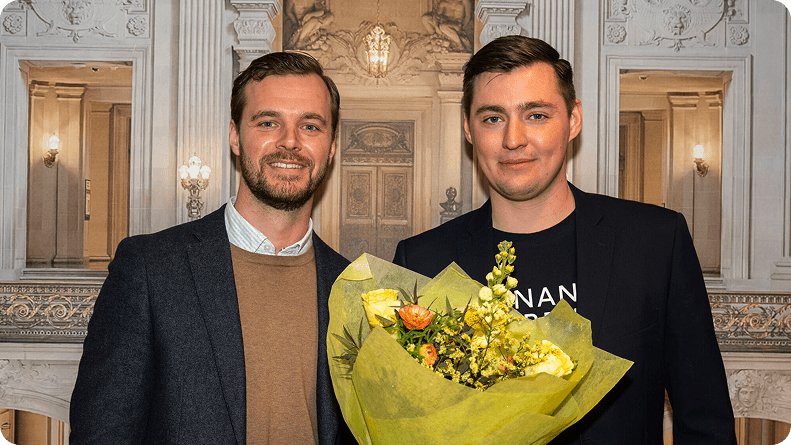 Two men smiling, one holding a bouquet of yellow and orange flowers, in an ornate hall.