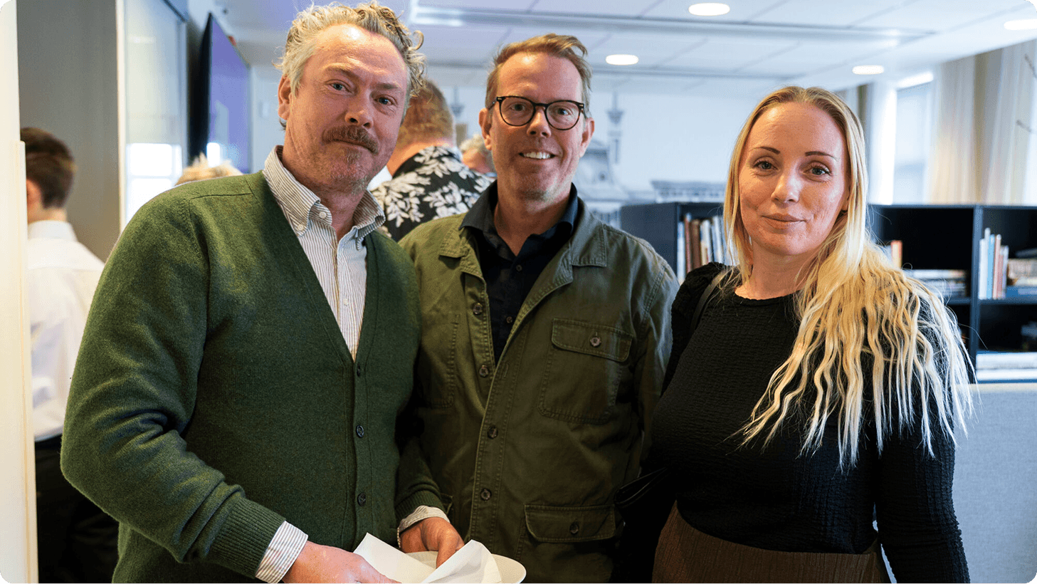 Three smiling colleagues, two men and one woman, stand together in an office environment.