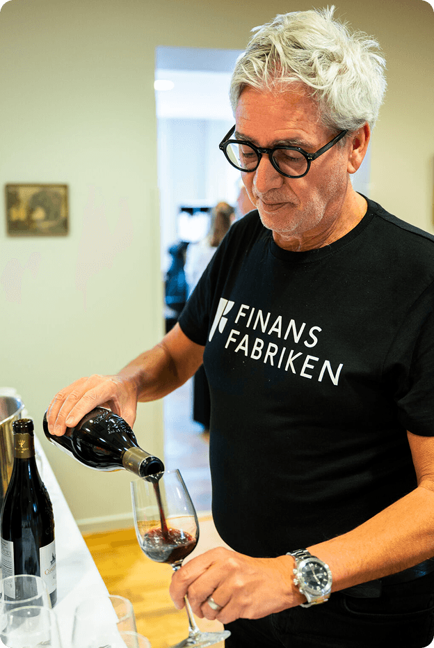 A man wearing glasses and a 'FINANS FABRIKEN' t-shirt pours red wine.