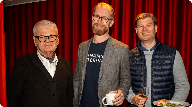 Three smiling men, one wearing a "Finans Fabrik" shirt, stand before a red curtain.