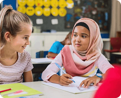 Two diverse schoolgirls collaborating happily at a desk in a vibrant classroom.
